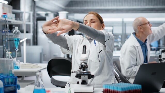 Histology technician examining pathology slides under microscope, fixing bad posture at workbench. Researcher driving medical innovation through cellular analysis work feeling back pain, camera B