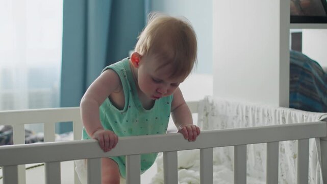 Infant girl leaning over crib railing and holding bars, learning to stand and control body balance in nursery room. Early childhood development at home