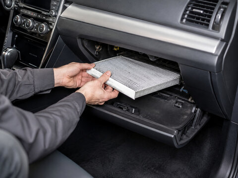 Mechanic or car owner hands replacing a dirty, dusty cabin air filter located behind the glove compartment dashboard box of a modern automobile interior. Vehicle maintenance service.