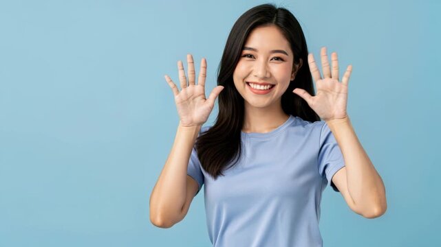 Smiling Woman Showing Ten Fingers: A young Asian woman with a bright smile and long dark hair holds up both hands, showing all ten fingers, against a calm blue background.