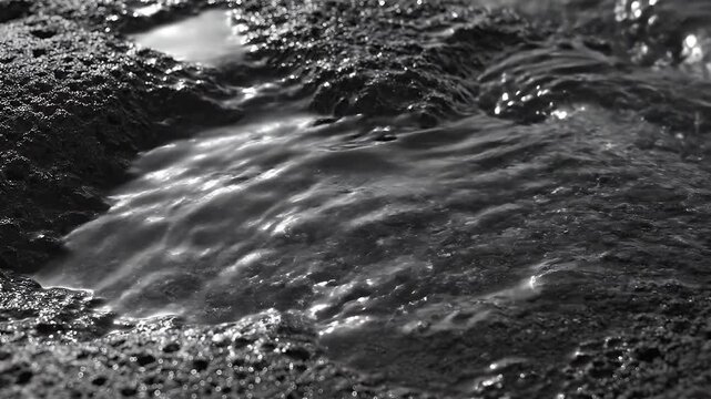 Black and white macro shot of water flowing over dark porous volcanic rock texture with silky motion blur and high contrast highlights, capturing the intricate detail of the basalt surface.