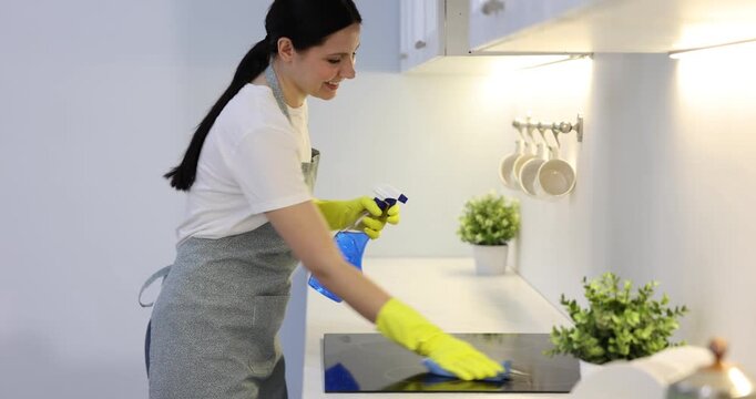 Spring cleaning. Smiling woman in apron and rubber gloves wiping electric stove with rag and bottle of detergent in kitchen