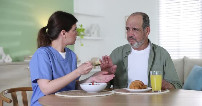 Caregiver with breakfast and elderly man refusing to eat it at wooden table indoors