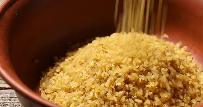 Raw bulgur pouring into bowl at wooden table, closeup