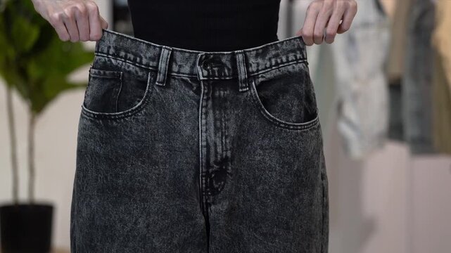 Slim woman lifts loose old black jeans to waist and lets them fall, falling at home. Concept of weight loss result, healthy lifestyle, wellness, body transformation, fitness progress and size change