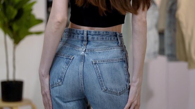 Young Caucasian woman in fitted blue high-waisted jeans turns gracefully and strokes hips in wardrobe home interior. Concept of flattering denim fit, feminine confidence, slim waist and casual style