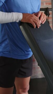 Vertical video: Resting hands on pad, male coach in blue top adjusting bench at gym, guiding client