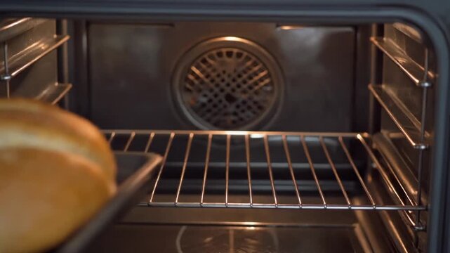 A golden-brown loaf of bread on a baking tray being carefully placed into a clean, preheated electric oven with a metal rack.