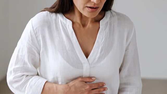 Young woman suffering from painful acid reflux and heartburn holding chest while standing indoors in white shirt