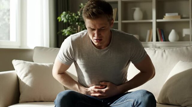 Young man sitting on a sofa in his living room experiencing severe stomach pain and acid reflux discomfort at home
