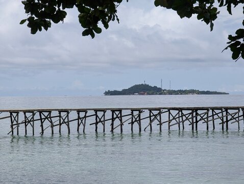 Wooden jetty extending across calm tropical waters toward Saonek Island in Raja Ampat, Indonesia, beneath dramatic cloudy skies, showcasing a serene coastal travel destination.