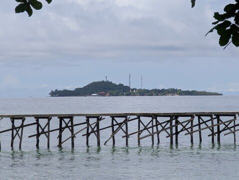 Wooden jetty extending across calm tropical waters toward Saonek Island in Raja Ampat, Indonesia, beneath dramatic cloudy skies, showcasing a serene coastal travel destination.
