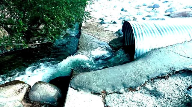 Water rushes from a large metal pipe into a rocky stream below with white foam and concrete edges surrounded by stones and green foliage in a natural outdoor setting
