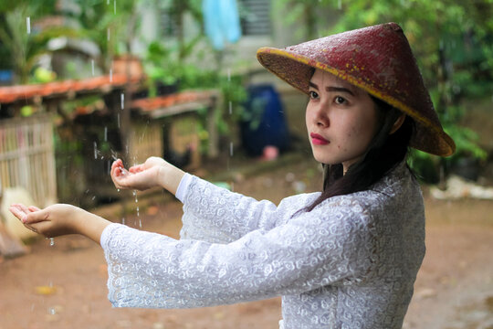 Indonesian village woman wearing traditional kebaya and conical bamboo hat standing under the rain, catching waterdrops.