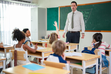 Elementary school teacher leading classroom discussion while students raise hands during interactive lesson in bright multicultural learning environment. © Wanwajee