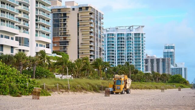Beach cleaning vehicle beside Miami oceanfront condominiums. Yellow beach cleaning vehicle on sand, palm trees and Miami Beach high-rise condos in background.