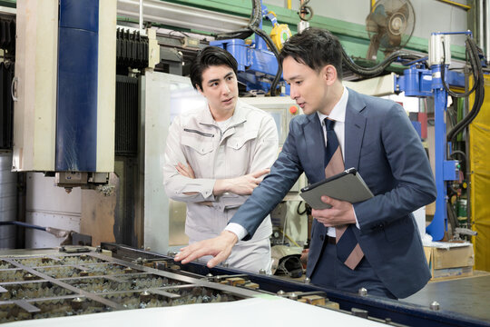 製造業の工場現場で作業着の技術者と営業が機械を見ながら見積もりや修理、メンテナンスの金額を悩む考えるのシーン