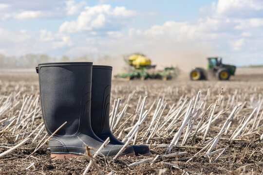 Spirit River, Alberta, Canada - May 17, 2026: John Deere 9450R tractor and 1910 commodity cart are seeding in the spring dusty field under blue sky, black gum boots are in the foreground in stubbles.