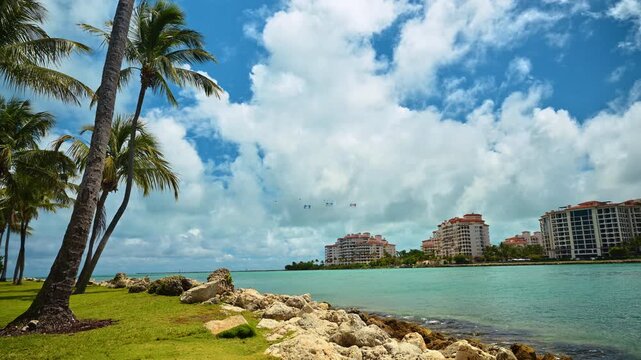 Fisher Island Condominiums Across Biscayne Bay In Miami. Palm trunks, rocks, turquoise bay water, and Fisher Island condos under cumulus clouds in Miami.