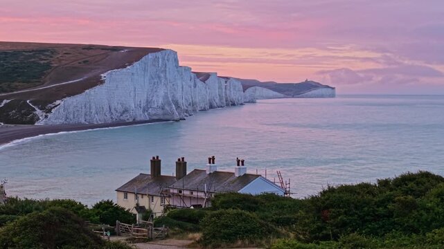 Aerial view of Seven Sisters Cliffs with a coastal cottage under a pink sunset sky in Exceat, England, United Kingdom.