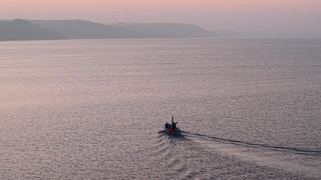 Aerial view of a small fishing boat sailing on calm water with rolling hills in the background during a soft pink sunset in Looe, England, United Kingdom.