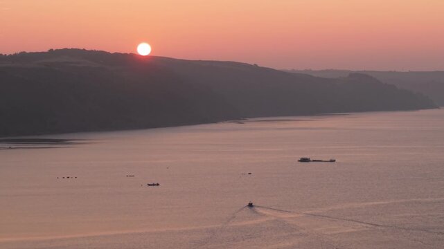 Aerial view of a small boat cruising on calm coastal waters during a vibrant sunrise over the rolling hills of the Cornish coast in Looe, England, United Kingdom.