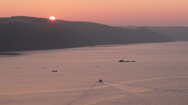 Aerial view of a small boat cruising on calm water with hills in the background during a pink sunset Looe, England, United Kingdom.