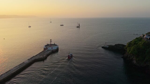 Aerial view of a small boat entering the harbor past a stone pier and lush green cliffs during a golden sunset in Looe, England, United Kingdom.