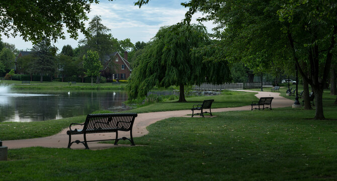 A winding asphalt walking path lined with park benches runs next to Culler Lake in Baker Park, Frederick, Maryland. Mature trees shade the green grass near neighborhood houses under a morning sky.