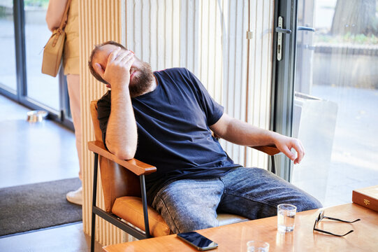 Tired white bearded man covering face and slumping in chair, clear signs of fatigue and frustration after long day, casual clothing and scattered phone on table underline stress and need for break