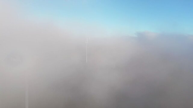 Aerial view of Windpark Freilanderalm wind turbines partially obscured by a thick layer of white clouds and mist under a bright blue sky in Pack, Steiermark, Austria.