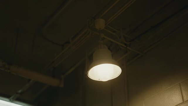 Warm industrial lamp casts soft light across a dim warehouse ceiling filled with pipes and wires, creating a moody atmosphere of solitude and quiet functionality