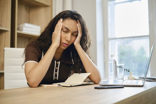 Tired young woman sits at her desk, visibly stressed and holding her head in her hands. The workspace features a notebook, a laptop, and a water bottle, with natural light coming from a window