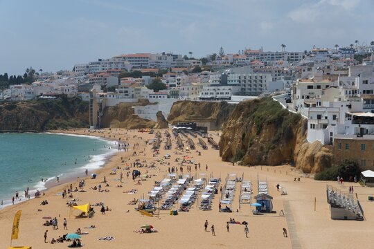Lively sandy beach with lots of people and sun loungers in front of picturesque cliffs and a coastal town, Albufeira, Faro, Algarve, Portugal