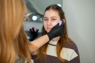 Naklejka premium Makeup artist applying foundation to a teenage girl's face with a sponge in a beauty salon. Professional makeup process, skincare, and modern beauty industry atmosphere.