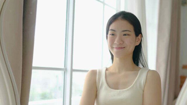 Happy young Asian woman touching her face looking in the mirror. Beautiful girl smiling with healthy clear glowing skin. Natural beauty, skincare, morning beauty routine and self care concept.