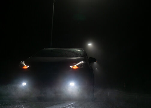 A dark car sits parked on a wet road at night. Its bright headlights, daytime running lights, and fog lights cut through the heavy mist.