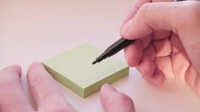 Hand Holding Black Pen Over Blank Block Of Yellow Sticky Notes Preparing To Write. Video footage of planning and preparing a note.