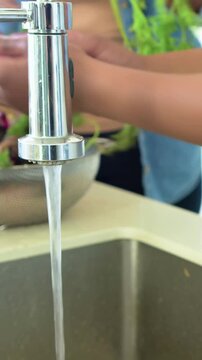 Vertical video: Washing cook in pink top scrubbing root under open tap, rinsing greens for cooking