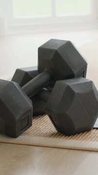 Vertical video: Cam dollying 3 black hex dumbbells on woven mat by window at home, showing home gym