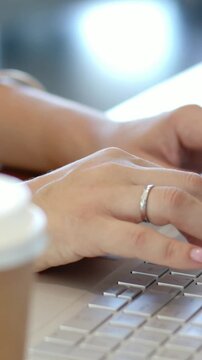Vertical video: Moving right hand to keys, ringed hands tapping laptop at desk while working, cup
