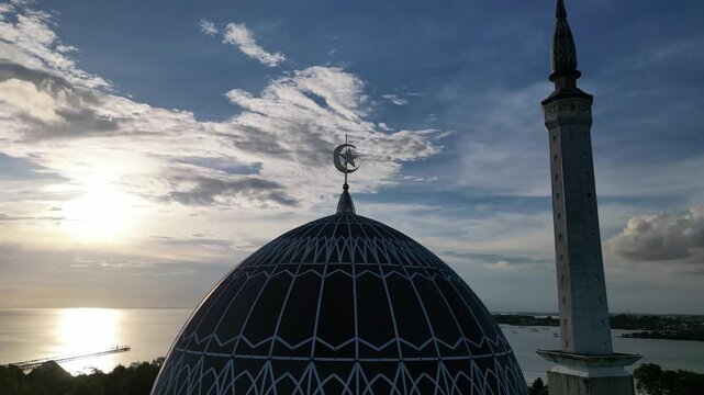 Aerial view of the dome and minaret of Masjid Raya Nur Ilahi overlooking the coast at sunset in Bukit Bestari, Riau Islands, Indonesia.