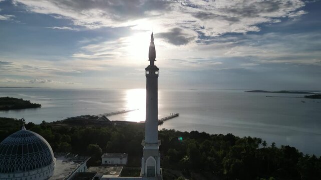 Aerial view of the tall minaret and dome of Masjid Raya Nur Ilahi overlooking the sea at sunset in Bukit Bestari, Riau Islands, Indonesia.