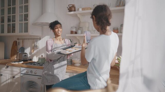Happy black woman holding baking sheet with cookies and posing for mobile photo. African american mother and daughter spending time together at home kitchen, motherhood and childhood, cook at home