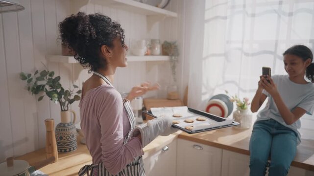 Teenage girl taking mobile photo of her mother with baking sheet in home kitchen . Happy african american family, single lady and her daughter cooking together in sunday morning, happy motherhood