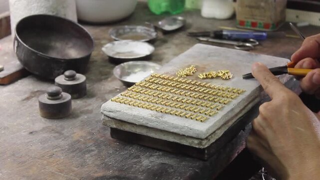 Macro shot of a jeweler's hand using tweezers to meticulously arrange small gold clasp links on a stone soldering block in a neat grid.