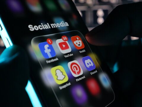 Closeup of a person holding a smartphone displaying a folder of popular social media apps including facebook, youtube, reddit, snapchat, pinterest, and discord