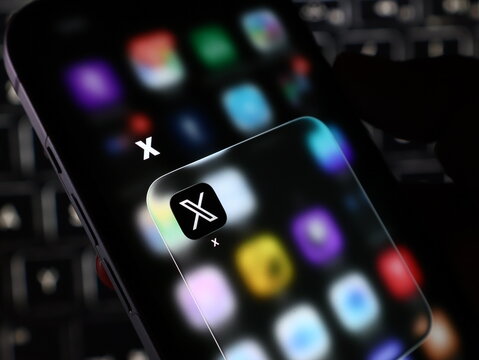 Closeup of a smartphone screen displaying the x app icon, formerly known as twitter, with a blurred background of app icons and a keyboard