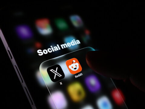 Closeup of a smartphone screen showing a social media folder with x and reddit app icons being touched by a finger in a dark room