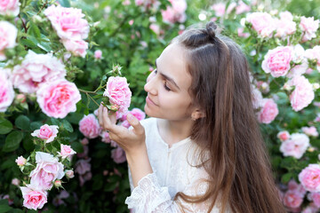Naklejka premium Beautiful girl smelling a pink rose while in a blooming garden of flowers. Romantic outdoor portrait with long hair, summer atmosphere. Charming lifestyle scene with nature, beauty, and tenderness.